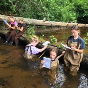 Girls in the midst of a stream collecting data on notebooks