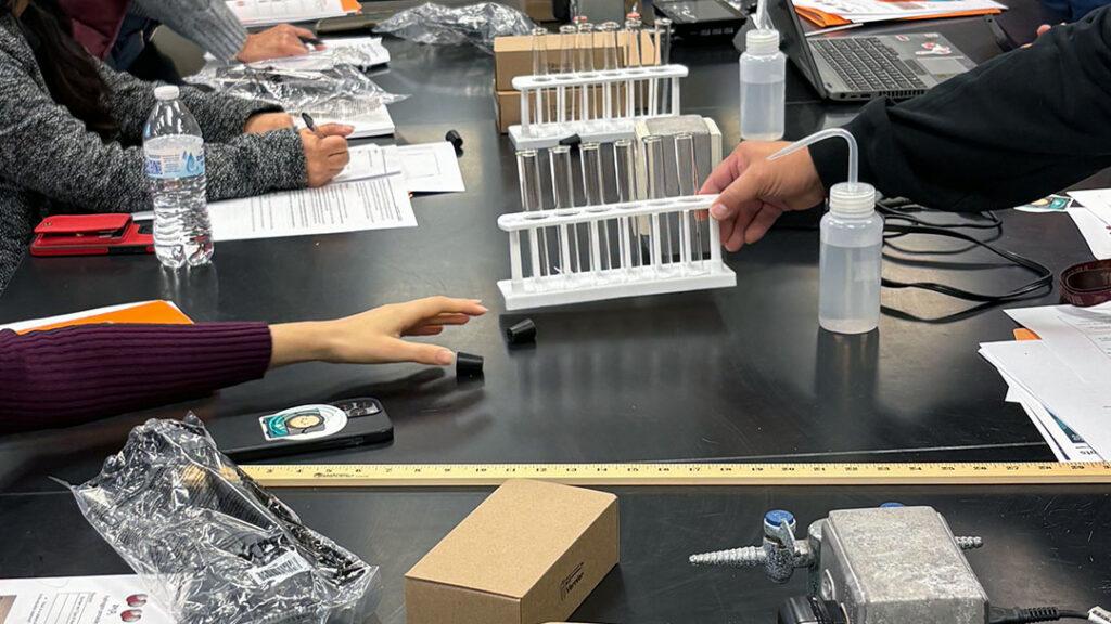 Instructors sit at a lab table with multiple pieces of science equipment, computers, and Vernier sensor boxes.