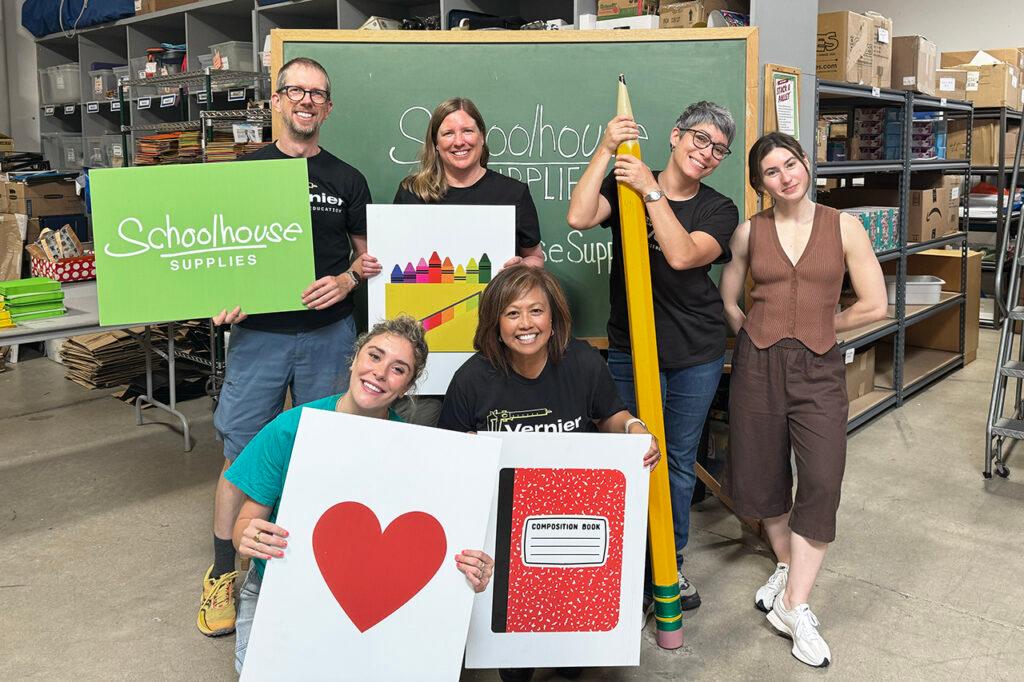 Vernier volunteers holding signs in the Schoolhouse Supplies warehouse; signs include illustrations of a box of crayons, a notebook, and a heart