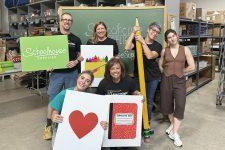 Vernier volunteers holding signs in the Schoolhouse Supplies warehouse; signs include illustrations of a box of crayons, a notebook, and a heart
